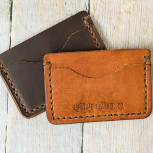 Puede incluir: Dos carteras de cuero, una marr&oacute;n y otra beige, con bordes cosidos. La cartera beige tiene el texto "LIFETIME LEATHER CO." grabado en relieve en la parte delantera.