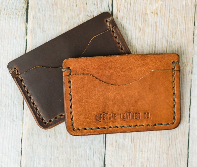 Puede incluir: Dos carteras de cuero, una marr&oacute;n y otra beige, con bordes cosidos. La cartera beige tiene el texto "LIFETIME LEATHER CO." grabado en relieve en la parte delantera.