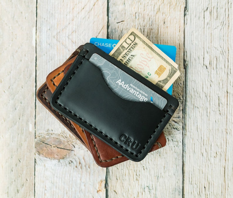 Puede incluir: Una cartera de cuero negro con una solapa de cuero marr&oacute;n y un detalle cosido. La cartera tiene una ranura para tarjetas y un bolsillo para efectivo. La cartera est&aacute; etiquetada como "CRU".