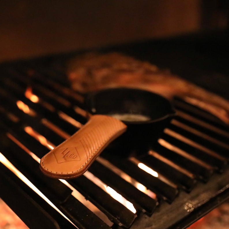 May include: A brown leather handle for a cast iron skillet, with the text "L.L.CO" embossed on the handle. The handle is sitting on a black grill grate with flames visible underneath.