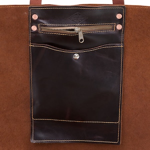 Könnte beinhalten: Eine braune Ledertasche mit einer dunkelbraunen Ledertasche und Reißverschluss. Die Tasche hat einen kleinen Metallknopf und die Tasche hat kupferfarbene Nieten.