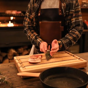May include: A person in a brown leather apron is preparing a piece of meat on a wooden cutting board. The board has three grooves running down the length of it. The person is using a knife to chop herbs over the meat.