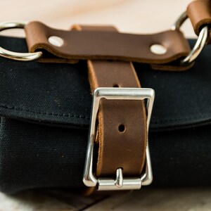 May include: Close-up of a black canvas roll with brown leather straps and silver metal hardware. The leather straps are secured with rivets and a silver buckle. The bag has a handle with a silver ring.