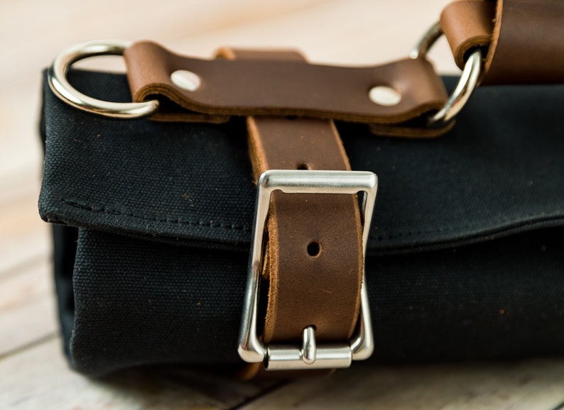 May include: Close-up of a black canvas roll with brown leather straps and silver metal hardware. The leather straps are secured with rivets and a silver buckle. The bag has a handle with a silver ring.