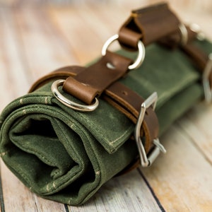 May include: Rolled-up green canvas tool roll with brown leather straps and silver metal buckles and rings. The canvas has a subtle speckled pattern. The leather straps are secured with metal buckles.