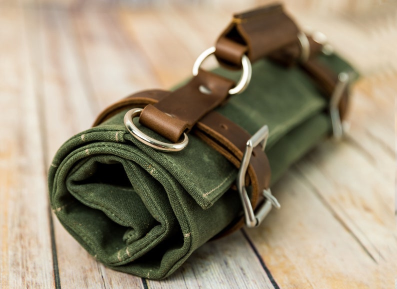 May include: Rolled-up green canvas tool roll with brown leather straps and silver metal buckles and rings. The canvas has a subtle speckled pattern. The leather straps are secured with metal buckles.