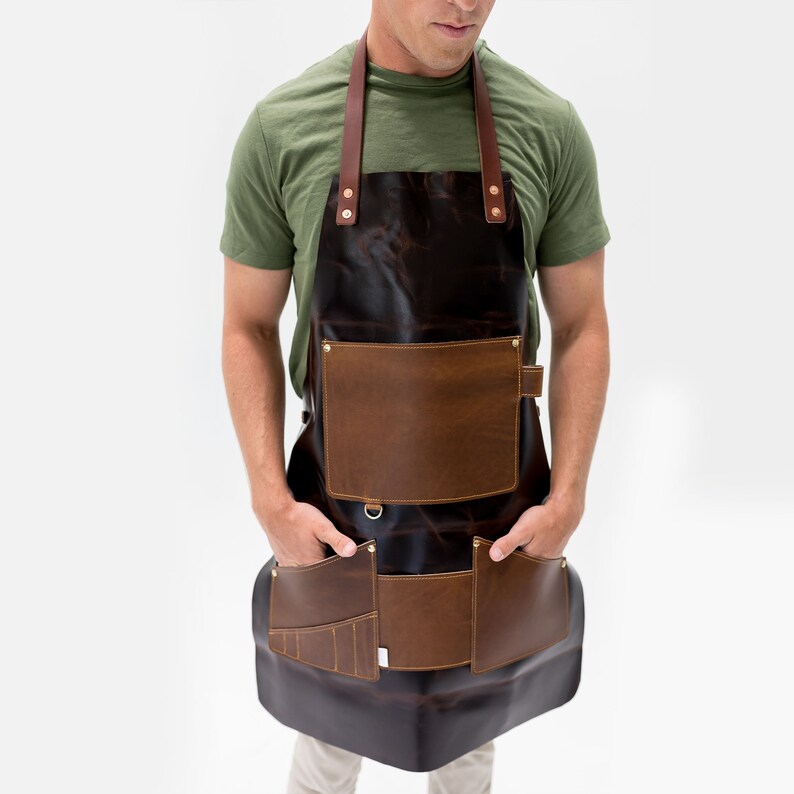 May include: A dark brown leather apron with two large front pockets and two smaller pockets. The apron has brown leather straps and a metal D-ring. The apron is worn by a person wearing a green t-shirt.
