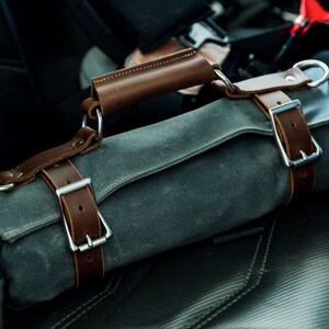 May include: A rolled-up, gray canvas tool roll with brown leather straps and handle. The roll is secured with two leather straps and silver buckles. The handle is also brown leather and attached with silver rings.
