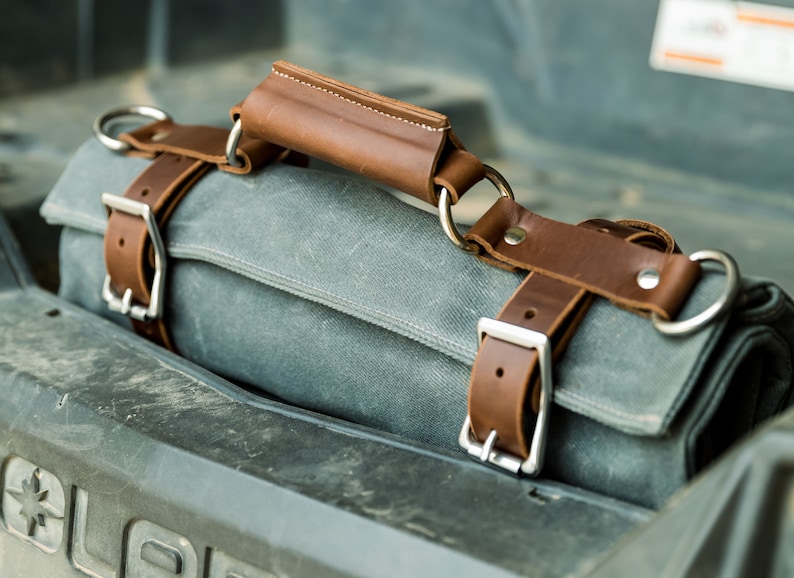 May include: A gray canvas tool roll with brown leather straps and handle. The roll is secured with silver buckles and metal rings. The handle is stitched with white thread.