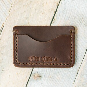 Puede incluir: Un porta tarjetas de cuero marr&oacute;n con un borde cosido y el texto "LIFETIME LEATHER CO." en relieve en la parte delantera.