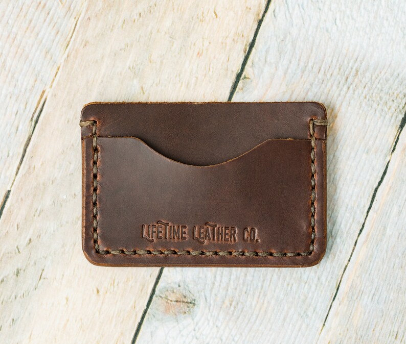 Puede incluir: Un porta tarjetas de cuero marr&oacute;n con un borde cosido y el texto "LIFETIME LEATHER CO." en relieve en la parte delantera.