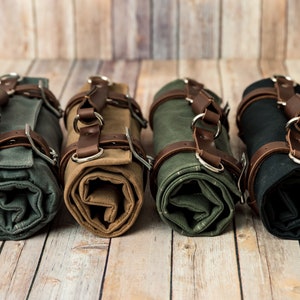May include: Four rolled canvas tool rolls in shades of gray, tan, olive green, and black. Each roll is secured with brown leather straps and silver metal rings. The rolls are displayed on a light brown wooden surface.