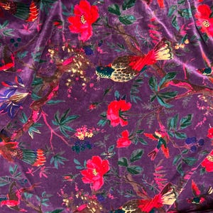 May include: A deep purple velvet fabric featuring a repeating pattern of vibrant red flowers, dark green leaves, and multi-colored birds with red, yellow, and blue plumage perched on brown branches throughout the textile surface.