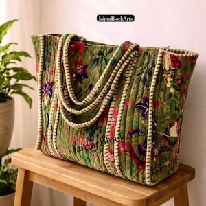May include: An olive green tote bag with a floral pattern in red, purple, and white. The bag has thick, cream-coloured rope handles and is on a wooden stool. The text "JaipurBlockArts" is visible on the bag.