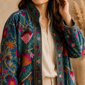 May include: A teal velvet jacket with colourful floral embroidery in pink, orange, and red. The jacket has a high collar and long sleeves, showcasing a bohemian style. The embroidery includes flowers and leaves.