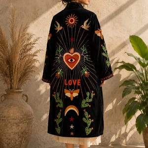 May include: A black velvet duster coat featuring colorful embroidery on the back. The design includes a central heart with an eye, the word LOVE, a sun, a moon, birds, moths, and cacti. The coat has long sleeves and a straight hemline.