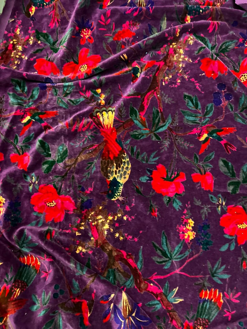 May include: A deep purple velvet fabric featuring a vibrant botanical print. The pattern displays bright red flowers, green leaves, and colorful birds perched on brown branches. The texture of the velvet creates subtle light and shadow effects.