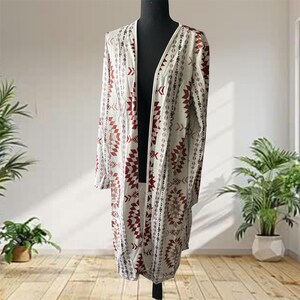 May include: A long-sleeved, open-front cardigan in a light cream color featuring a repeating geometric pattern in shades of deep red and dark brown. The garment has a straight hemline and falls to approximately knee length on the display form.