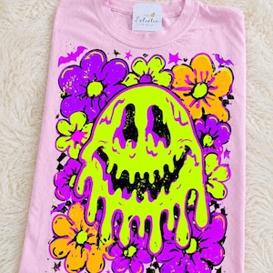 May include: Pink t-shirt featuring a neon green, smiling ghost design with dripping details, surrounded by purple and yellow flowers. The shirt also has a small tag that says "The Eclectic Peach".