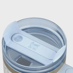 May include: Close-up of a floral-patterned tumbler with a light blue lid. The lid features a sliding mechanism and a logo of a winged creature. The tumbler has a gold band and a floral design on the lower portion.