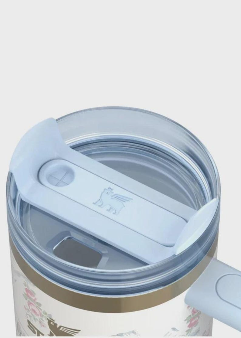 May include: Close-up of a floral-patterned tumbler with a light blue lid. The lid features a sliding mechanism and a logo of a winged creature. The tumbler has a gold band and a floral design on the lower portion.