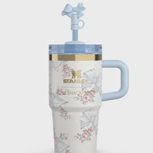 May include: A white Stanley travel mug with a floral and bow pattern. It features a light blue handle, lid, and straw with a bow detail. The text "STANLEY" and "LOVESHACKFANCY" are printed on the front.