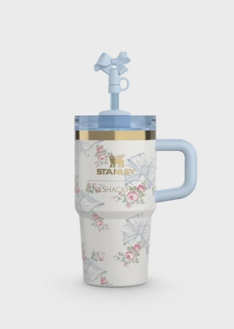 May include: A white Stanley travel mug with a floral and bow pattern. It features a light blue handle, lid, and straw with a bow detail. The text "STANLEY" and "LOVESHACKFANCY" are printed on the front.