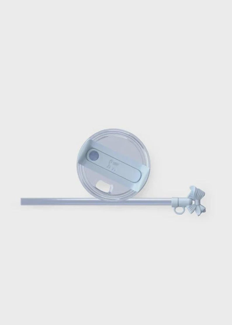 May include: A pale blue tumbler lid with a matching straw and decorative bow. The lid features a sliding closure and a small, embossed design. The straw is straight and the bow is detailed.