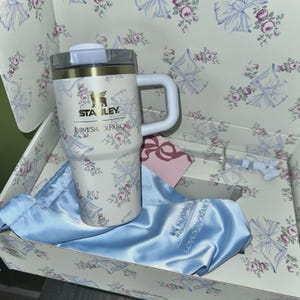 May include: A white Stanley travel mug with a floral and bow pattern, a white handle, and a clear lid. The mug is in a box with a matching floral design, a blue satin bag, and a pink tag. The text "LOVESHACKFANCY" is visible.