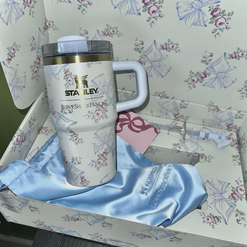 May include: A white Stanley travel mug with a floral and bow pattern, a white handle, and a clear lid. The mug is in a box with a matching floral design, a blue satin bag, and a pink tag. The text "LOVESHACKFANCY" is visible.