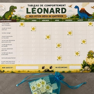 May include: A rectangular behavior chart titled TABLEAU DE COMPORTEMENT LÉONARD featuring dinosaur illustrations, a weekly grid with French text, and yellow star stickers. A blue organza drawstring bag filled with additional star stickers is below.