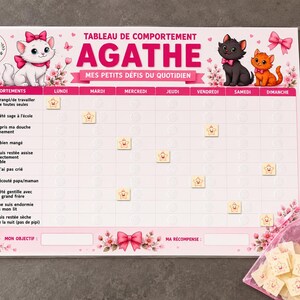 May include: A rectangular behavior chart titled TABLEAU DE COMPORTEMENT AGATHE MES PETITS DÉFIS DU QUOTIDIEN featuring illustrations of three kittens, pink bows, and cherry blossoms. The grid includes daily columns and rows with icons for tasks, accompanied by a pink organza bag filled with small square star tokens.