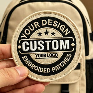 May include: A circular embroidered patch featuring a beige background with black text and borders. The text reads YOUR DESIGN, CUSTOM, YOUR LOGO, and EMBROIDED PATCHES, with three black stars centred above the word CUSTOM in a white rectangular frame.