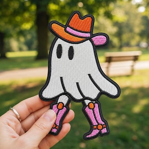 May include: An embroidered patch featuring a white ghost character wearing an orange cowboy hat with a pink band. The ghost has two black oval eyes and is wearing pink cowboy boots with orange accents, all outlined in a thick black border.