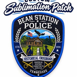 May include: A shield-shaped patch with a blue border featuring the text CUSTOM SUBLIMATION PATCH at the top. The center displays BEAN STATION POLICE, a log cabin, an American flag, a bald eagle, two figures with rifles, and HISTORICAL CROSSROADS.