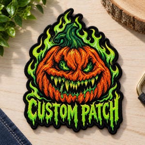 May include: An embroidered patch featuring an orange jack-o'-lantern with a menacing expression, sharp yellow teeth, and green glowing eyes. The pumpkin is surrounded by vibrant green flames, and the text CUSTOM PATCH is stitched in green.