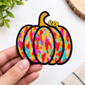May include: An embroidered pumpkin patch featuring a vibrant, multi-coloured abstract pattern of yellow, pink, red, and light blue shapes. The pumpkin has a black stitched border, a yellow stem, and a small yellow curly vine detail on the top right.