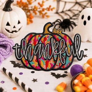 May include: An embroidered patch shaped like a pumpkin with a multicoloured abstract pattern in shades of pink, yellow, and teal. The word thankful is stitched across the centre in white thread with a thick black outline and border.