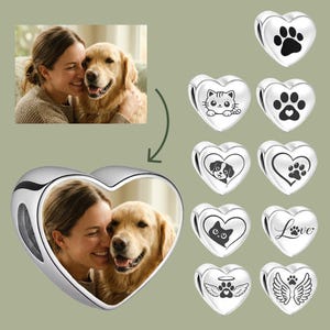 Op de afbeelding: Een zilveren hartvormige bedel met een aangepaste kleurenfoto van een persoon en een golden retriever. Rechts worden acht extra zilveren hartvormige bedels getoond, elk gegraveerd met verschillende ontwerpen en iconen met een huisdierthema.