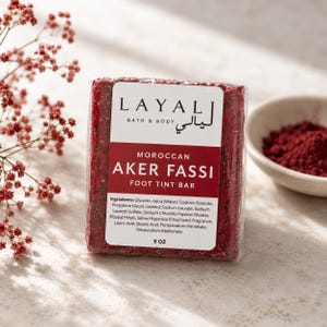 May include: A rectangular Layal Bath & Body Moroccan Aker Fassi foot tint bar in a deep red color with visible seeds. The white label features the brand name, product name, and a list of ingredients, with a net weight of 5 oz printed at the bottom.