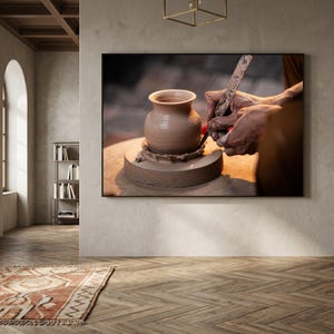 May include: A framed photograph of a potter at work, shaping a clay vase on a pottery wheel. The potter's hands are visible, using tools to refine the vase's form. The vase is light brown, and the background is blurred.