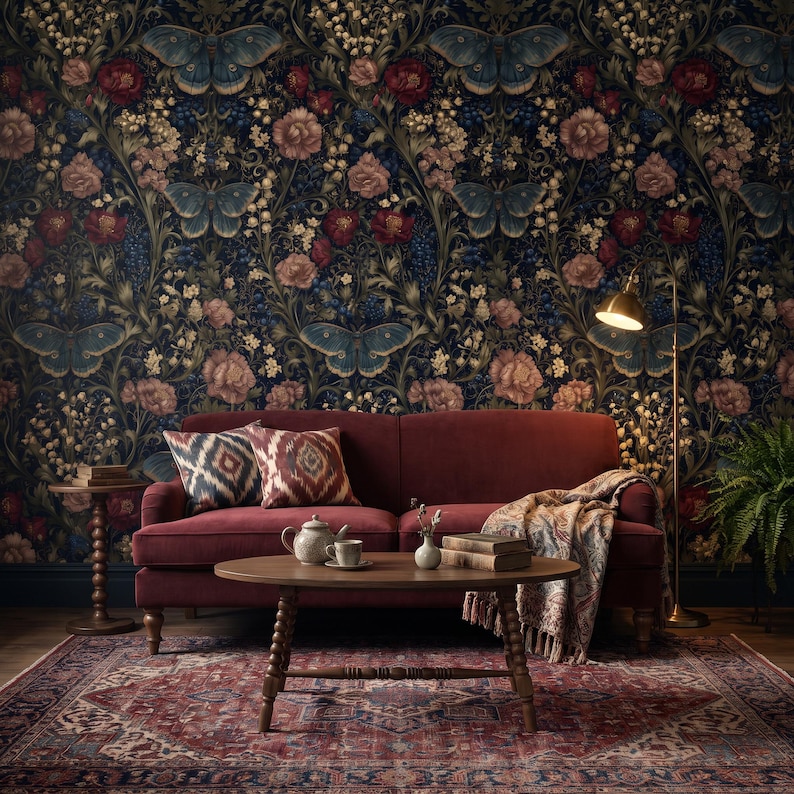 May include: A deep red velvet sofa with two patterned throw pillows and a draped blanket, positioned in front of dark floral wallpaper featuring blue butterflies. A wooden coffee table holds a teapot, cup, and books, with a floor lamp nearby.