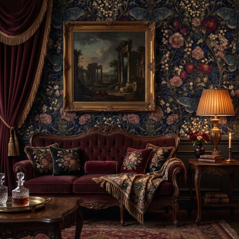 May include: A deep burgundy velvet sofa with tufted backrest, adorned with floral-patterned throw pillows and a fringed tapestry throw blanket. The sofa is positioned beneath a framed landscape painting and beside a table lamp with a pleated shade.