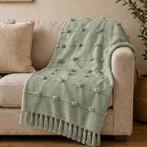 May include: A sage green throw blanket featuring a textured woven pattern with horizontal rows of small raised tufted knots and a decorative fringe trim along the bottom edge, draped over the arm and seat of a neutral colored sofa.