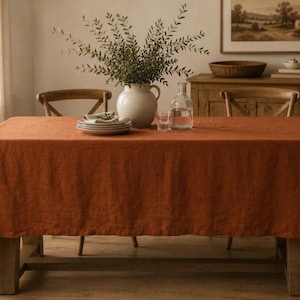 Pode incluir: Uma toalha de mesa retangular de cor laranja queimado sólido sobre uma mesa de jantar de madeira. O tecido tem uma textura tecida visível. Sobre a mesa, há um jarro de cerâmica branca com folhagem verde, uma pilha de pratos brancos e um copo.