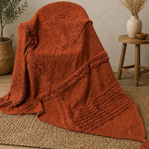 May include: A burnt orange throw blanket draped over a chair, featuring a textured surface with raised tufted geometric patterns, including diamond shapes and horizontal lines, finished with a single tassel at the bottom corner of the fabric.
