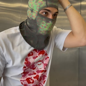 Könnte beinhalten: Graue Sturmhaube mit grünem und rosa Muster, kombiniert mit einem weißen T-Shirt mit rosa Camouflage-Grafik. Das T-Shirt hat ein rundes Logo unten links.