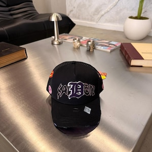 Puede incluir: Una gorra tipo trucker negra con parte trasera de malla y visera curva con detalles desgastados. El panel frontal muestra el texto "SADBOY" en una fuente estilizada de color morado y blanco, junto con pequeños parches circulares en los laterales.