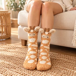 May include: A pair of fuzzy, plush crew socks featuring a giraffe pattern in shades of tan and cream. The socks include three-dimensional giraffe ears and small brown horns at the top cuff, with embroidered eyes and a snout on the front.