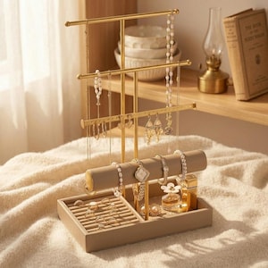 May include: A gold-toned metal jewelry organizer stand with a beige rectangular base and a cylindrical bar for bracelets. The stand features multiple tiers for hanging necklaces and earrings, with various pieces of jewelry displayed on the hooks.